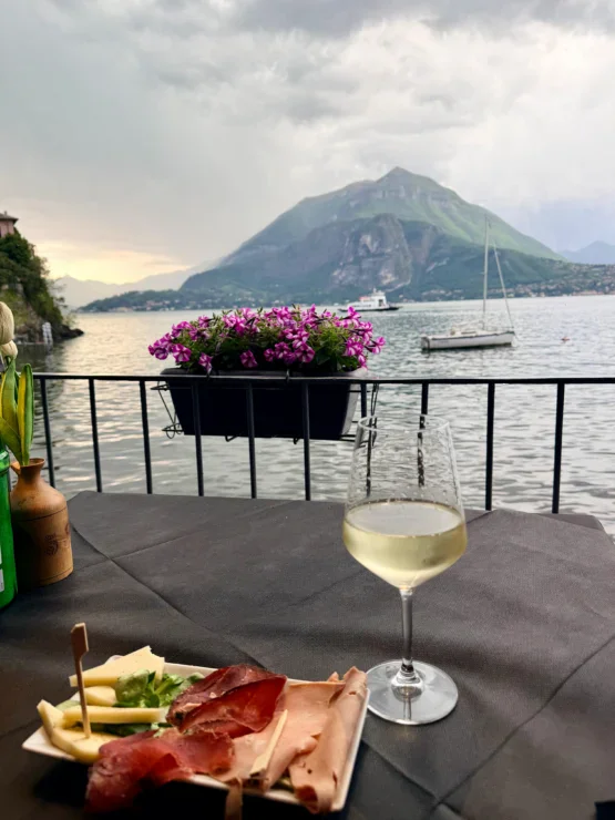 A glass of white wine and appetizers on a table overlook a lake with boats and a large green mountain in the background.
