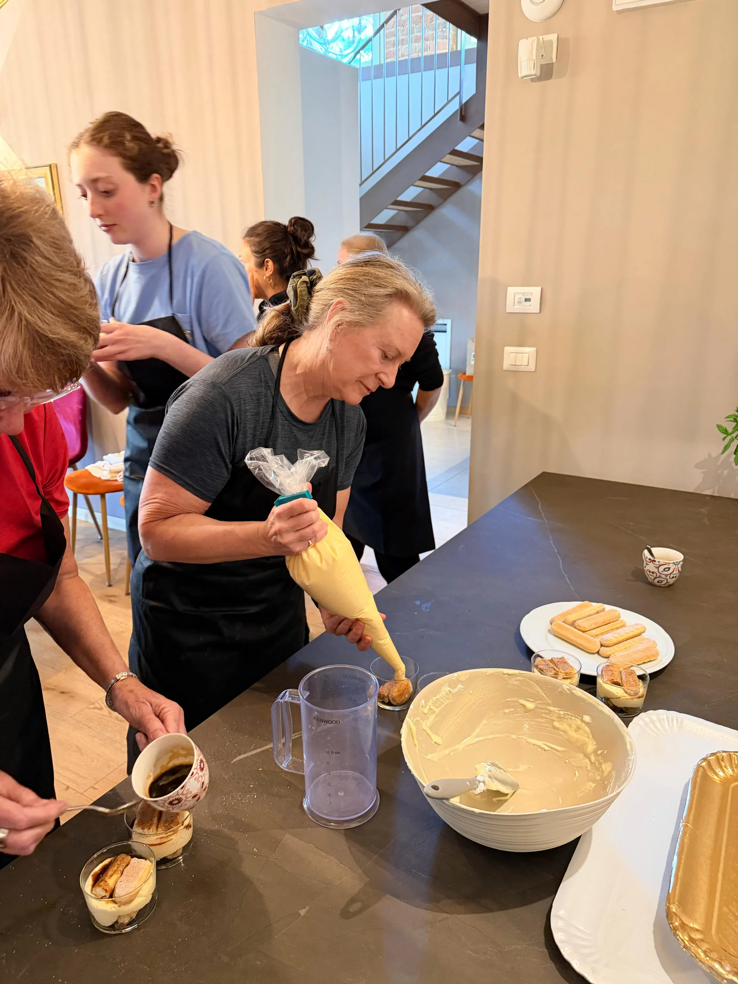 Several people stand around a kitchen counter assembling desserts, with one woman piping cream into cups and others preparing ingredients.