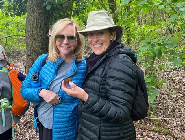Two women in puffer jackets stand in a wooded area, smiling at the camera; one holds an object in her hand.