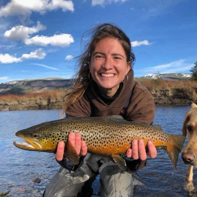 Person holding a large brown trout while kneeling in a river. A dog is partially visible beside them. Fishing net in the foreground.