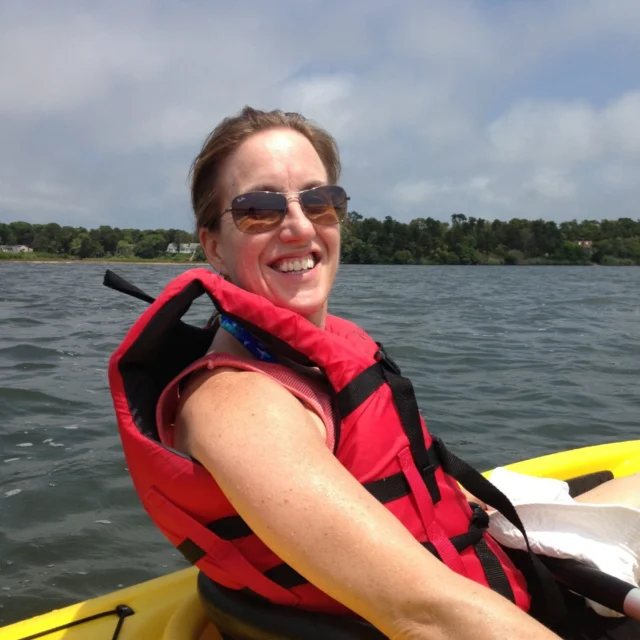 A person wearing sunglasses and a red life jacket smiles while sitting in a yellow kayak on a lake.