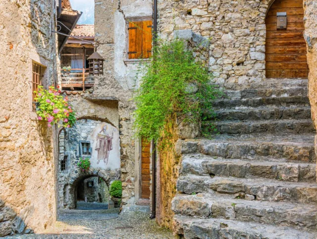 Stone alleyway in an old village, featuring a mural, wooden doors, stone steps, and flowering plants.