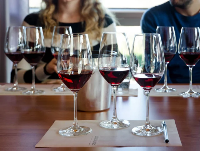 Two people sitting at a table with several glasses of red wine and a pen on a paper mat in front of them.