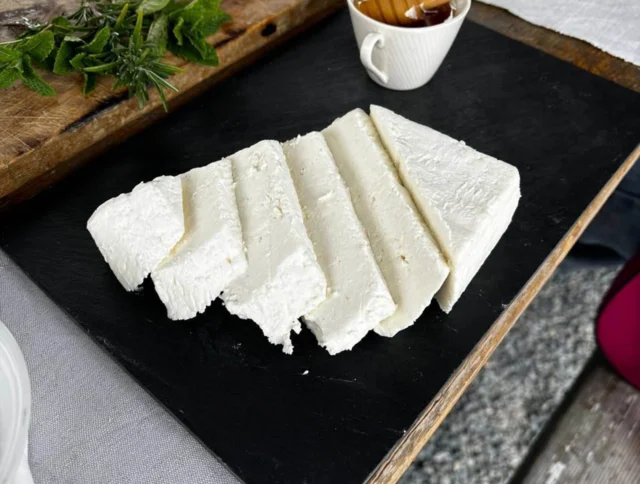 Slices of cheese on a black tray with a cup of honey. Nearby, there's a bowl of salad and some herbs on a wooden board.