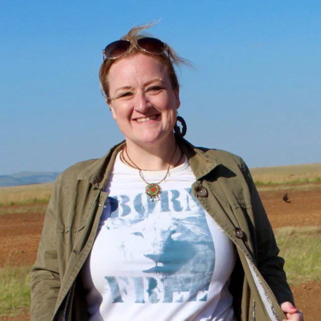 A person standing outdoors, smiling, wearing sunglasses on their head, a green jacket, and a "Born Free" t-shirt with a scenic field in the background.