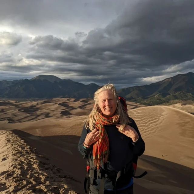 A person stands on a sand dune, wearing a colorful scarf and backpack. The background features a vast desert landscape under a cloudy sky, with mountains in the distance.