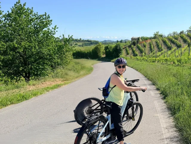 Person wearing a helmet and sunglasses stands with a bicycle on a sunny, winding country road lined with greenery and vineyards.