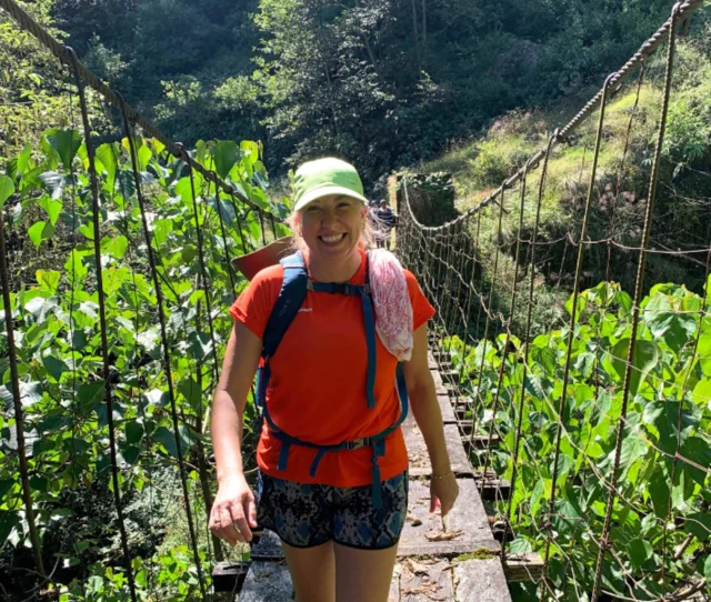 Person wearing an orange shirt and green cap walks on a wooden suspension bridge surrounded by lush greenery.