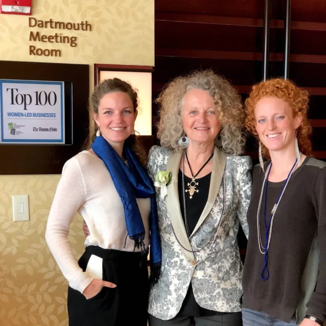 Three people standing together indoors, smiling. One person wears a floral jacket, another a blue scarf, and the third a necklace. A sign reads "Dartmouth Meeting Room" above them.