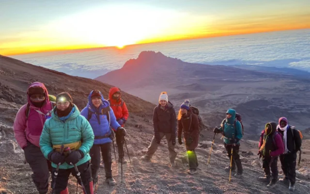 A group of hikers in winter gear stands on a mountain slope at sunrise, with a distant peak and a blanket of clouds in the background.