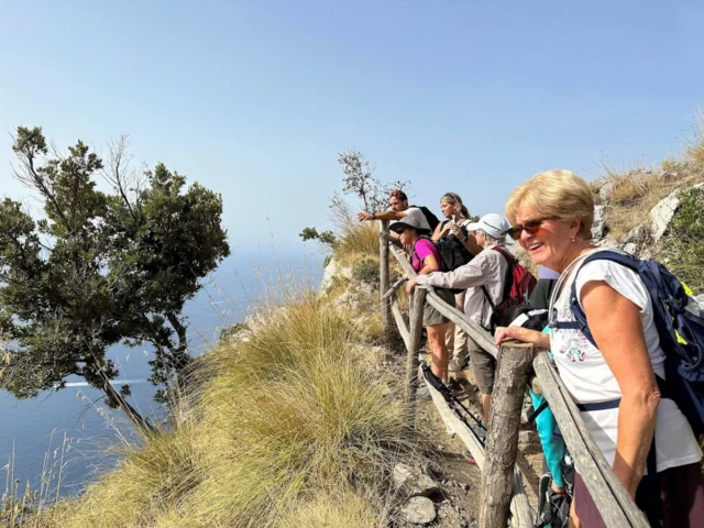 A group of hikers stands on a scenic cliffside trail overlooking the sea, with some leaning on a wooden fence amid dry grass and shrubs.