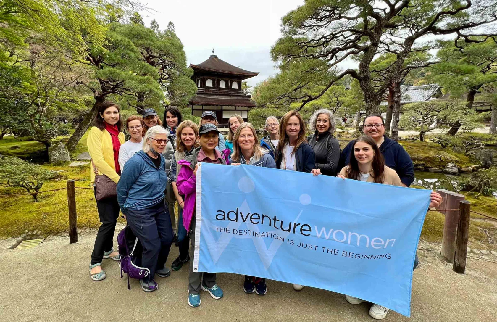 A group of women stand outdoors holding a banner that reads "adventurewomen." Trees and a wooden structure are in the background.