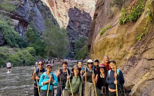 A group of hikers stand in a rocky riverbed, surrounded by steep canyon walls and lush greenery under a clear blue sky.