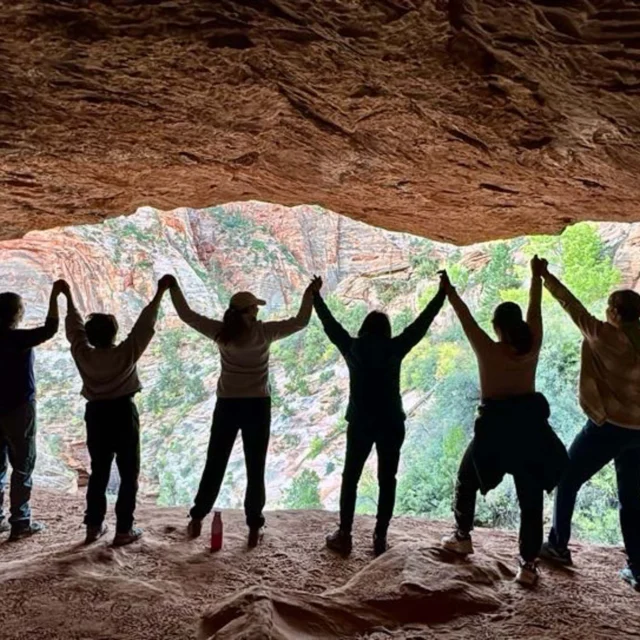 Silhouetted group of people holding hands under a rocky overhang with a scenic view of cliffs and greenery in the background.