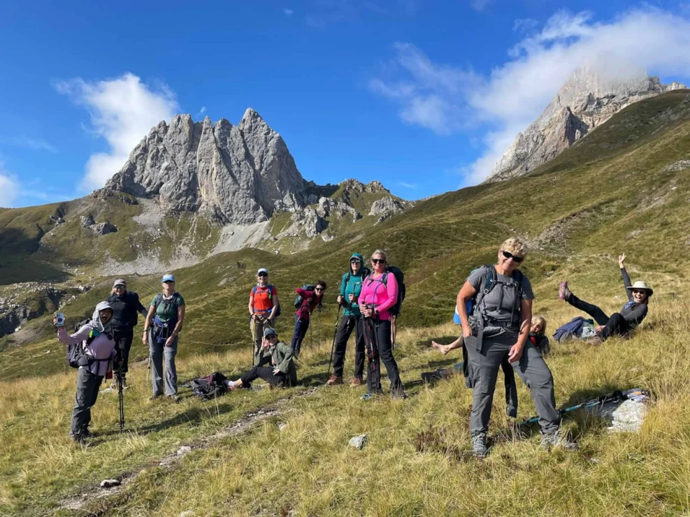 A group of hikers with backpacks and trekking poles stand and sit on a grassy slope with rocky peaks in the background under a clear blue sky.