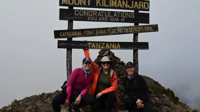 Three people pose in front of a Mount Kilimanjaro sign indicating they are at Cathedral Point Shira Peak, Tanzania.