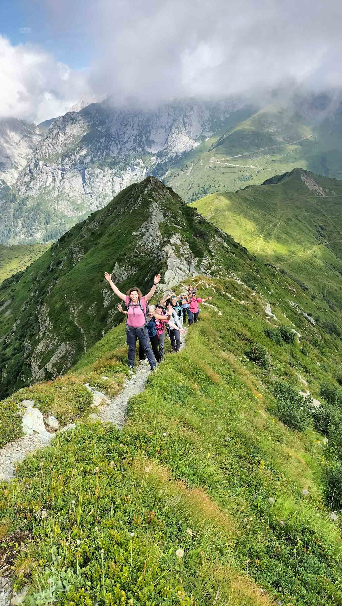 A group of people hiking on a narrow mountain trail surrounded by lush green hills and cloudy skies.