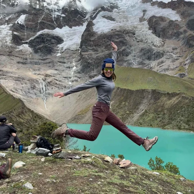 Person jumping joyfully in front of a turquoise lake and snow-capped mountains, while others sit nearby.