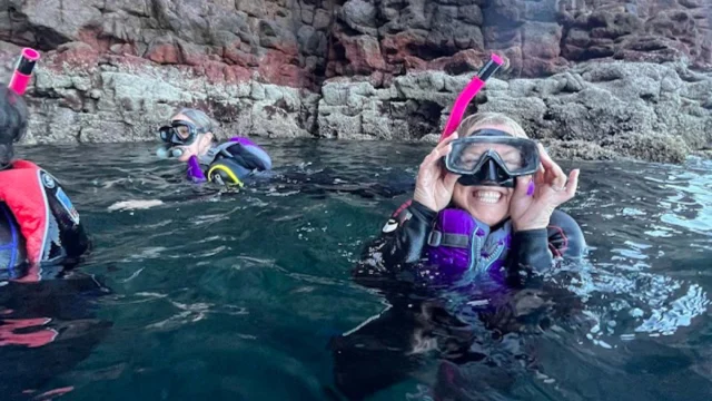 Three people wearing wetsuits, goggles, and snorkels are in the water near rocky cliffs.