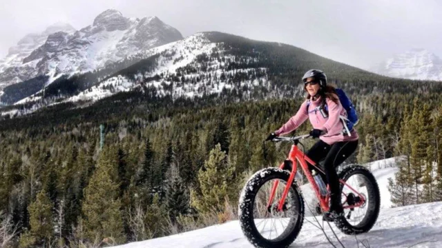 A person on a fat tire bike rides through a snowy landscape with forested hills and mountains in the background.