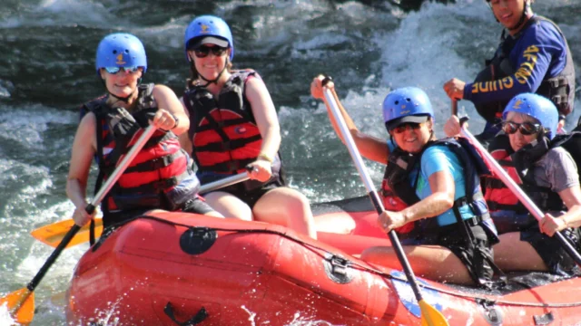 A group of people wearing helmets and life vests are whitewater rafting in a red inflatable raft on a river, guided by a person in a blue jacket.