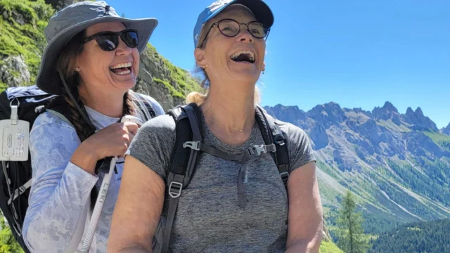 Two people wearing hats and backpacks smiling and laughing on a mountain trail with clear skies and mountains in the background.