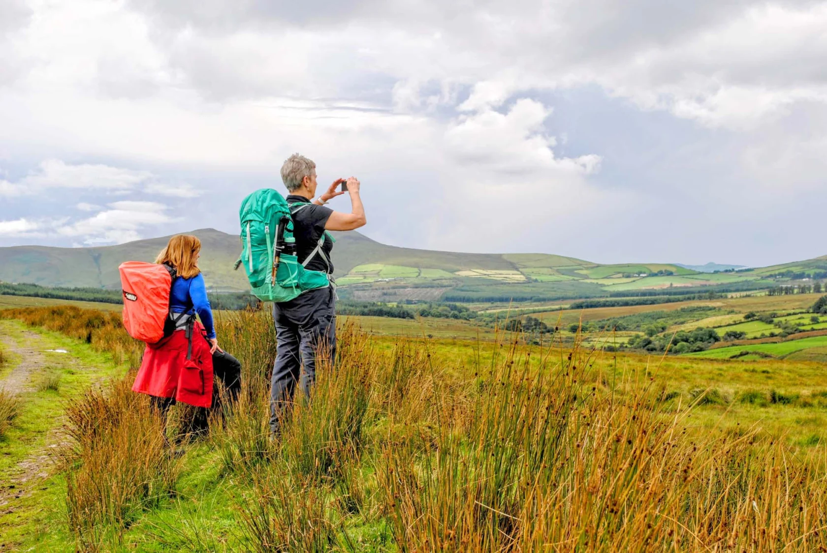 Two hikers with backpacks stand on a grassy hill, looking at a landscape view of rolling hills and fields under a partly cloudy sky.