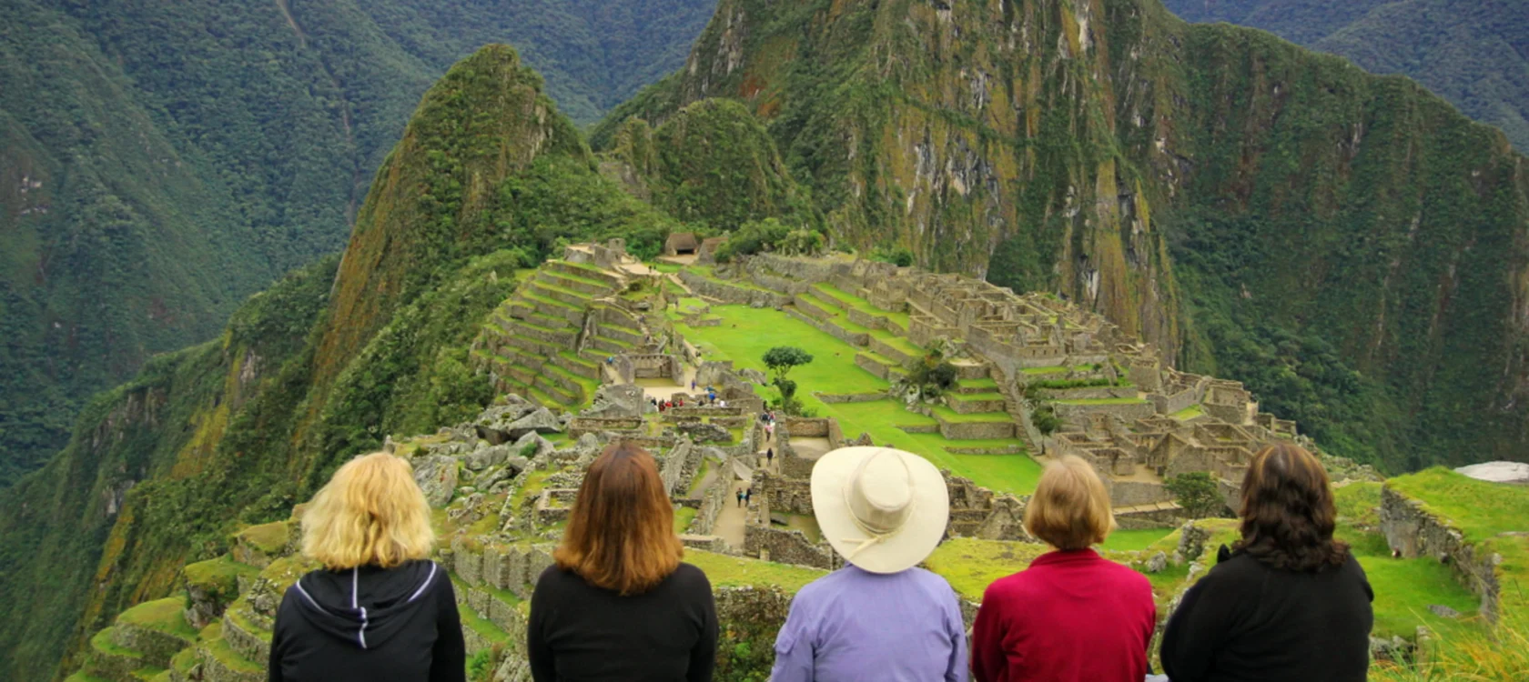 Five people sit facing the ancient Incan city of Machu Picchu on a green mountaintop, with steep, lush mountains in the background.