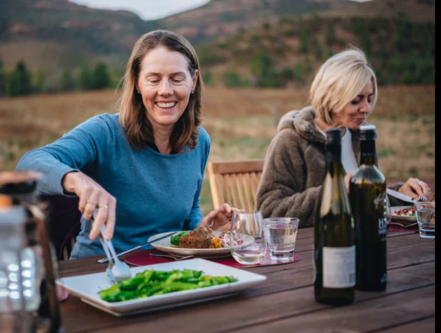 Two women enjoy an outdoor meal at a wooden table with bottles and plates of food in a scenic countryside setting.