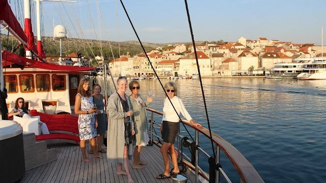 Five women on a boat deck holding glasses, with a coastal town and calm water in the background.