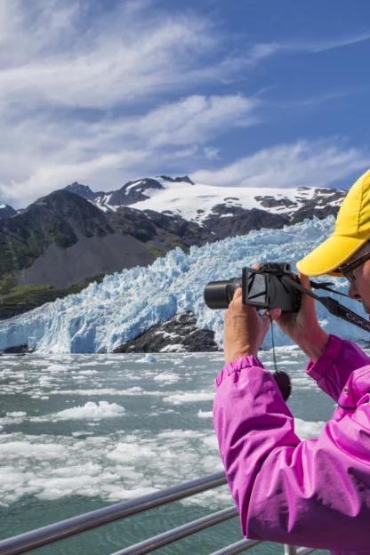 A person in a yellow cap and pink jacket takes a photo of a glacier and icy water from a boat on a sunny day.