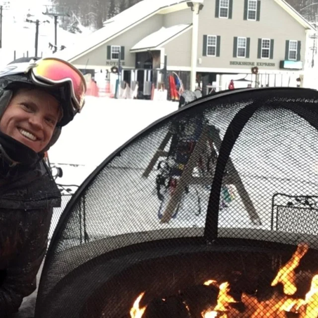 Person in winter gear smiles beside a fire pit at a snowy ski resort with ski lifts and a lodge in the background.