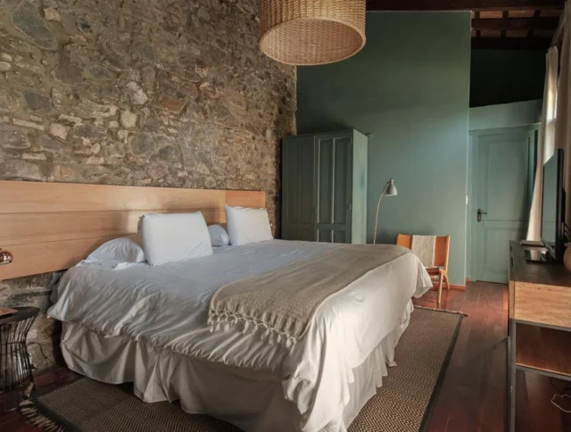 A bedroom with a stone accent wall, green painted walls, a double bed with white bedding, a wicker lampshade, and a wooden chair.