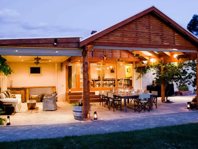 Outdoor covered patio with seating area and dining table, surrounded by greenery. It features warm lighting and a wooden ceiling.