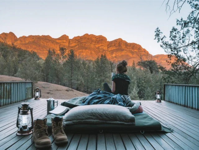 Person sitting on a deck with a sleeping bag and boots, overlooking a scenic mountain range during sunset.