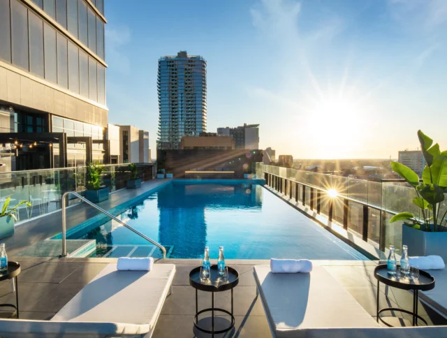 Rooftop pool with lounge chairs and tables, overlooking city buildings at sunrise.