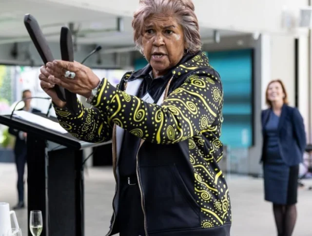 Elderly person in patterned jacket speaks while holding objects at a podium. Another person stands in the background.