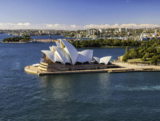 Aerial view of the Sydney Opera House with its distinctive sail-like architecture, surrounded by water, and the cityscape in the background under a clear blue sky.