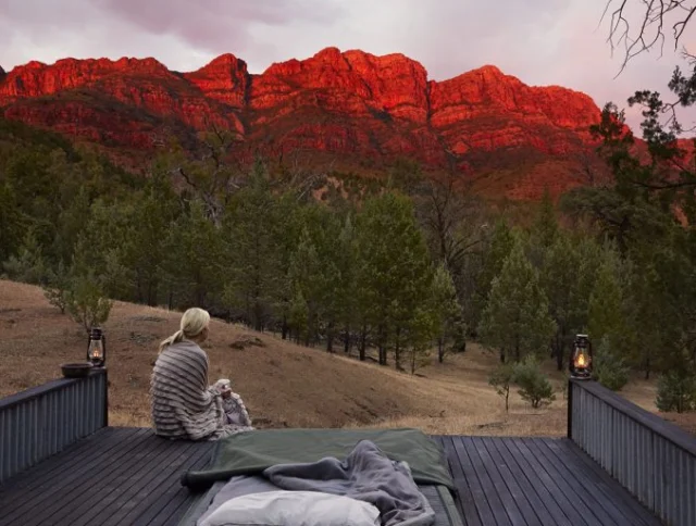 Person sitting on a wooden deck wrapped in a blanket, facing a mountainous landscape at sunset with a red-orange glow. Lanterns are placed on the deck.