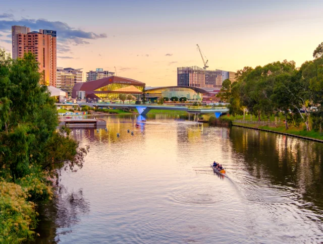 Rowers on a river at sunset with modern buildings and a lit bridge in the background.