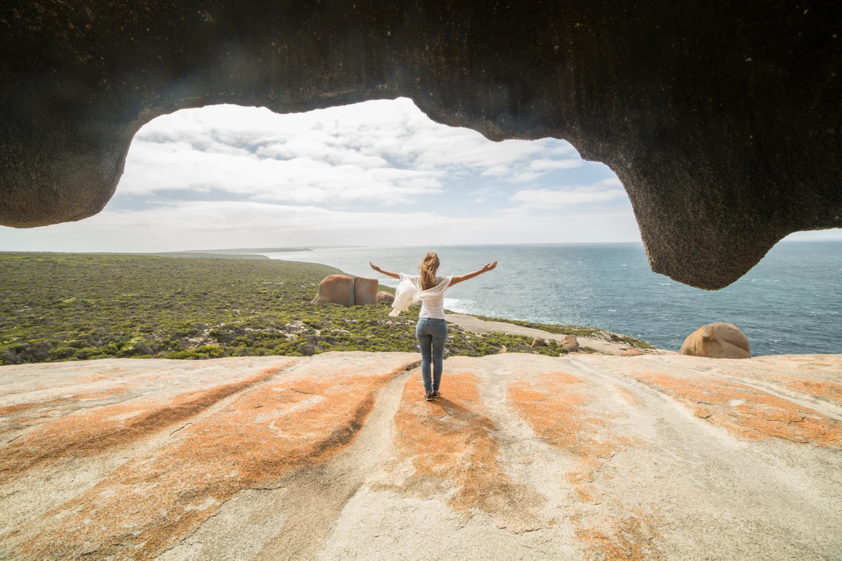 Person with arms outstretched stands on a rocky cliff overlooking the ocean under a rocky arch formation with a cloudy sky.