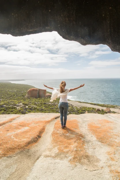 Person with arms outstretched stands on a rocky cliff overlooking the ocean under a rocky arch formation with a cloudy sky.