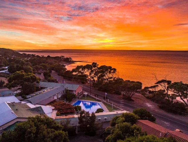 Sunset over a coastal town with a pool in foreground, trees and buildings nearby, and an orange sky reflected on the water.