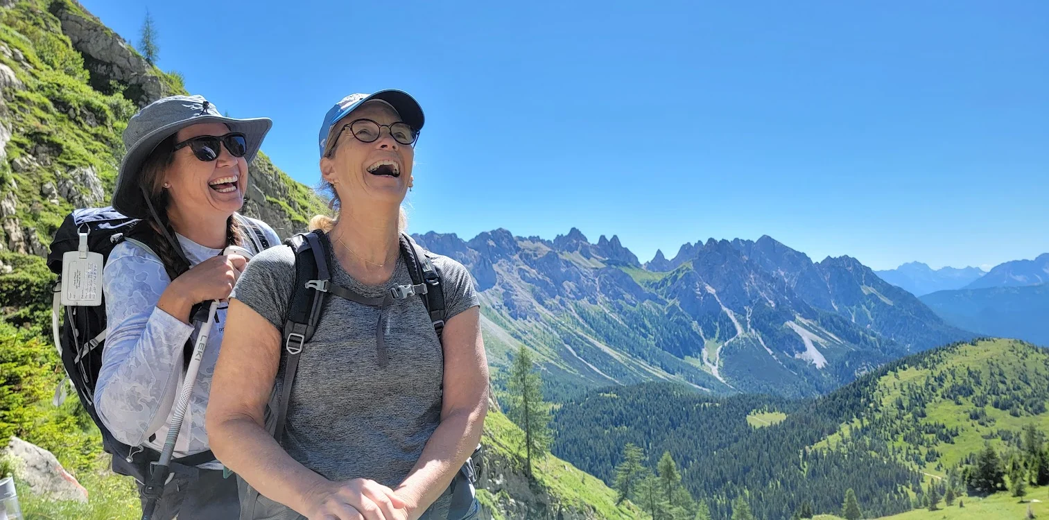 Two women with backpacks laugh while hiking on a sunny day in a mountainous landscape.