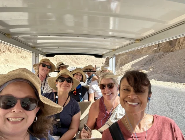 A group of people wearing hats and sunglasses smiles while seated in an open tram on a sunny day.