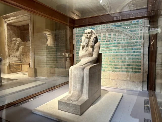 Stone statue of a seated figure in a glass display case, with a tiled wall in the background, inside a museum setting.