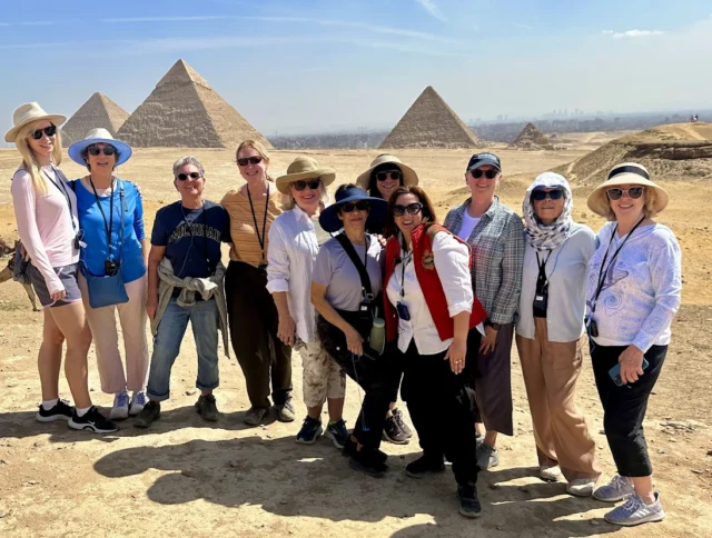 A group of people stand in front of the Pyramids of Giza on a sunny day, wearing casual clothing and hats.