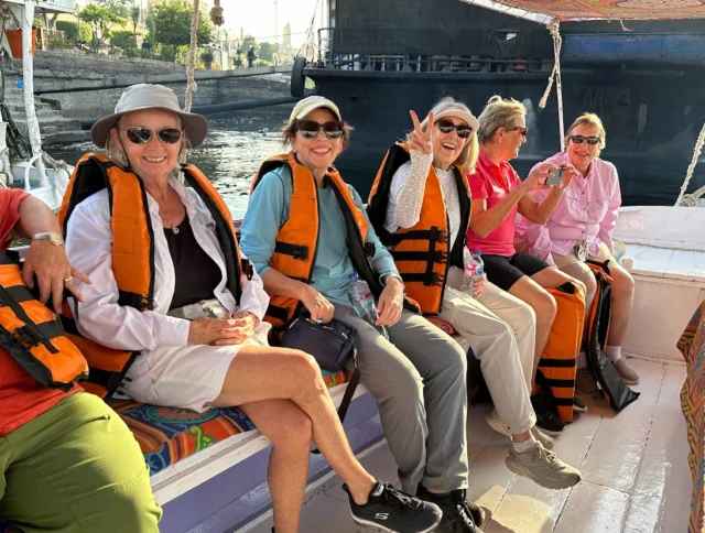 A group of five women wearing life jackets sit on a boat, smiling and posing for a photo in the sunlight.