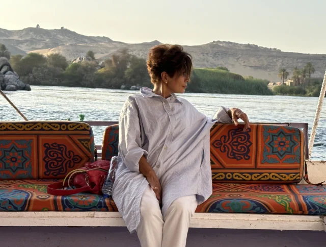 Person in a long shirt and white pants sits on a patterned boat bench, overlooking a river with hills and trees in the background.