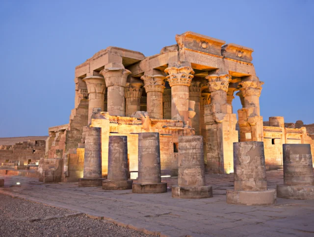 The image shows the ancient Kom Ombo Temple in Egypt, illuminated at dusk. The temple features large stone columns and intricate carvings.